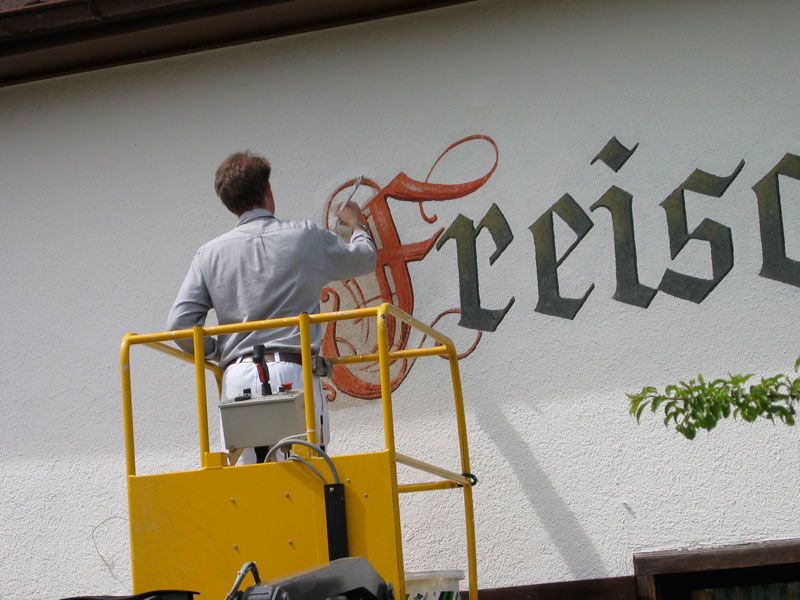 Maler auf einer Hebebühne malt einen Schriftzug an eine Fassade